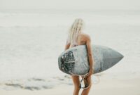 An adult woman carrying a surfboard walks towards the ocean on a sunny day in Bali, Indonesia.